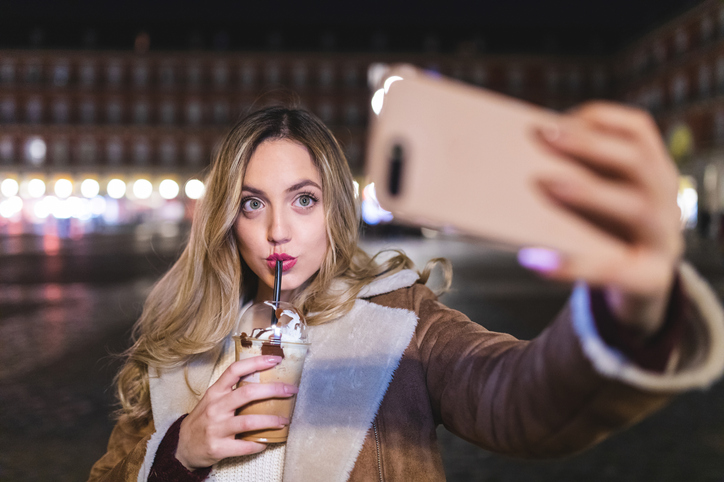Young woman drinking frappe coffee and taking selfie on city street at night, Madrid, Spain