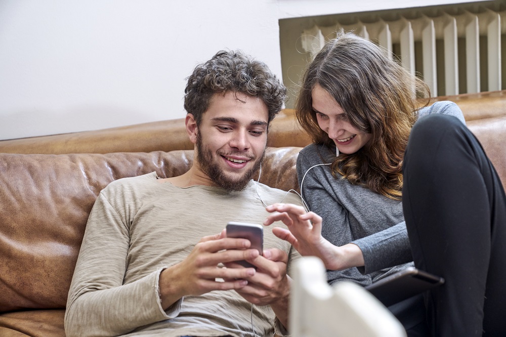 Young man and young woman sitting on couch sharing cell phone and earphones
