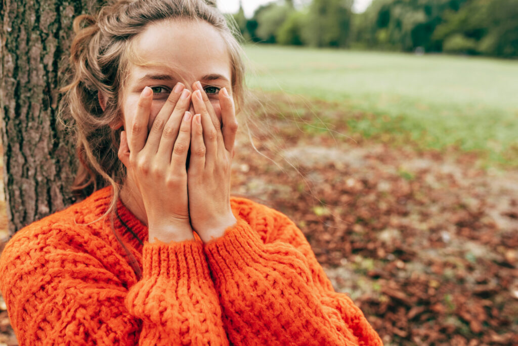 Horizontal candid portrait of a happy young woman smiling, hiding her face with two hands, playing peek a boo game in the park. Female wearing an orange sweater has joyful expression outdoors.