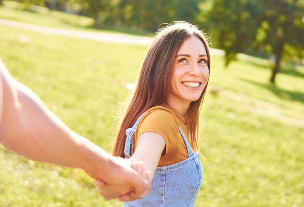 Young happy couple walking together in a park and holding hands – Girlfriend looking back to her boyfriend and smiling to him – Friends having fun together – Outdoor recreation lifestyle concept