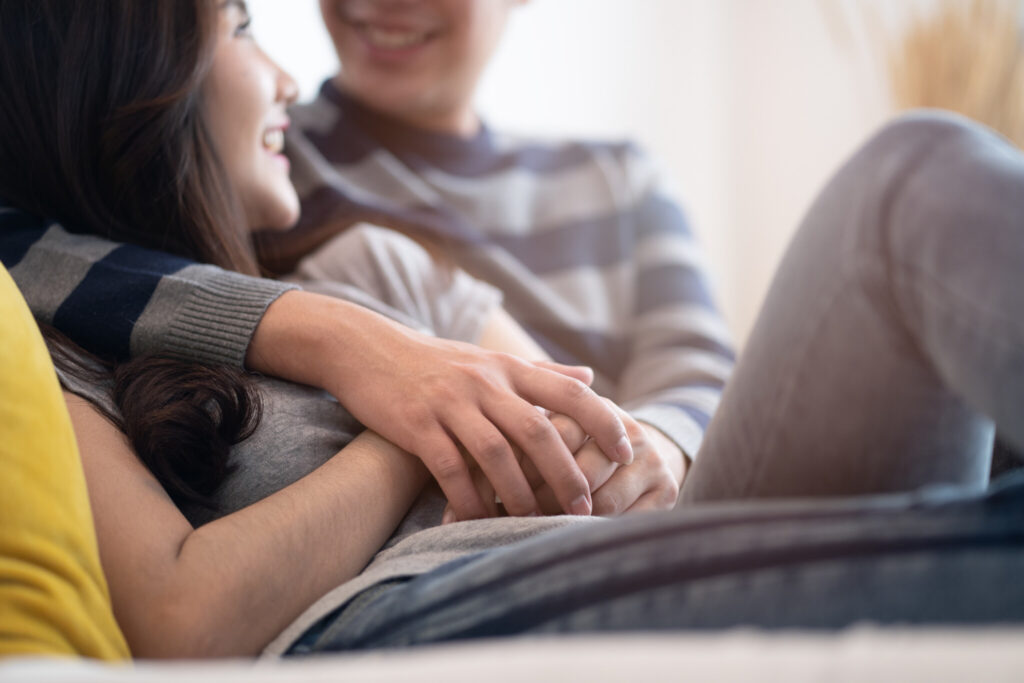 Lovely Asian couple sitting on bed in bedroom talking together with happiness. Boy hugging girl and holding hand, be hand in hand. Crop shot focus on hands. Man looking at woman eyes and smile face.