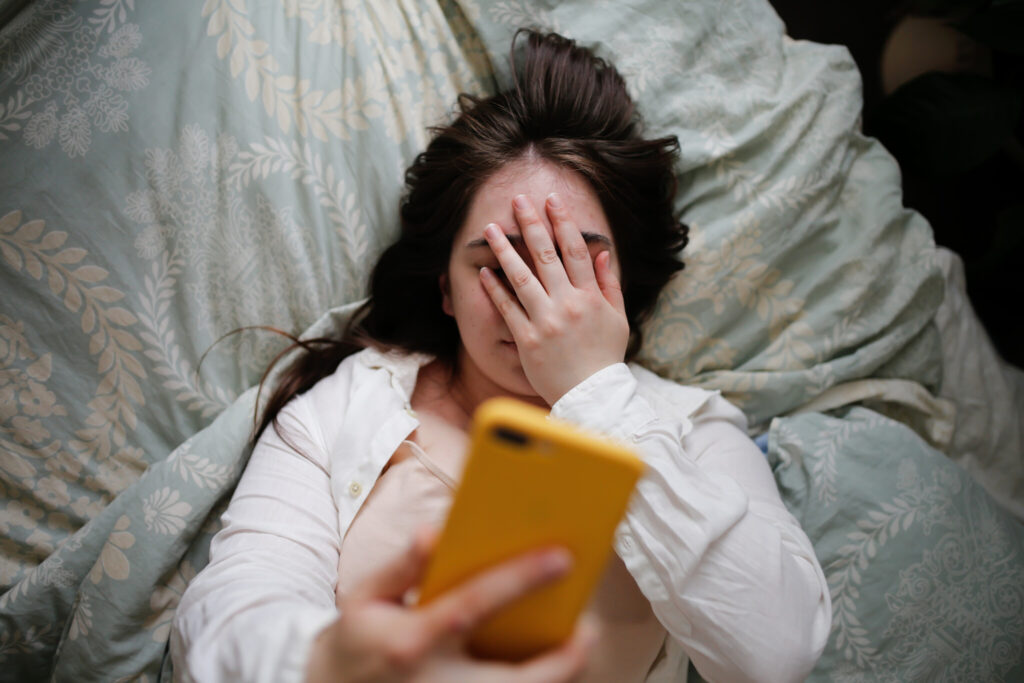 Sleepy overweight young woman with flowing hair using phone on bed, close face palm. Top view