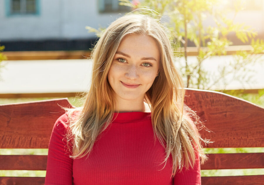 Outdoor portrait of pretty woman with light hair, freckles and blue eyes sitting on wooden bench in open air waiting for her best friend to come. Woman with appealing appearance posing outdoors