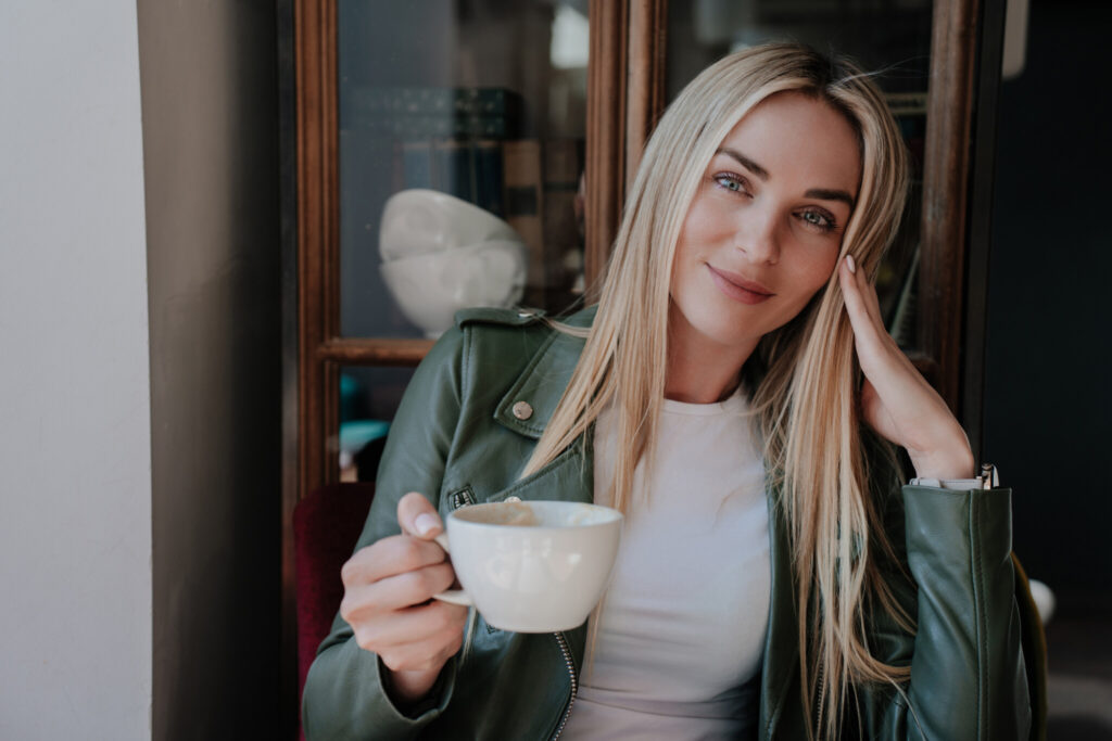 Adorable blonde girl in leather jacket and white t-shirt sitting at cafe holding a cup of coffee, looking at camera with cute smile. Pretty caucasian model having break while shoots. Leisure and youth