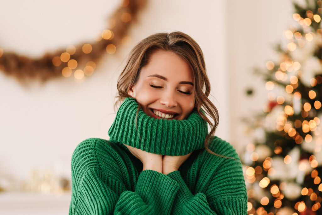 Portrait of a funny emotional cheerful young woman with horns on her head in a green cozy warm sweater laughing having fun holding and eating a lollipop and ginger cookies on Christmas day on the background of a decorated Christmas tree at home in winter