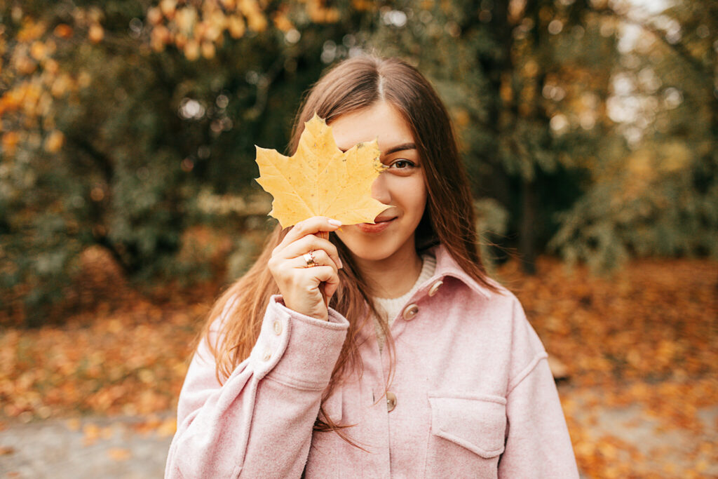 Positive brown-haired woman with long straight hair dressed in cozy autumn clothes is having fun in front of camera, posing with leaf on her face inautumn park. Indian summer. Weekend.