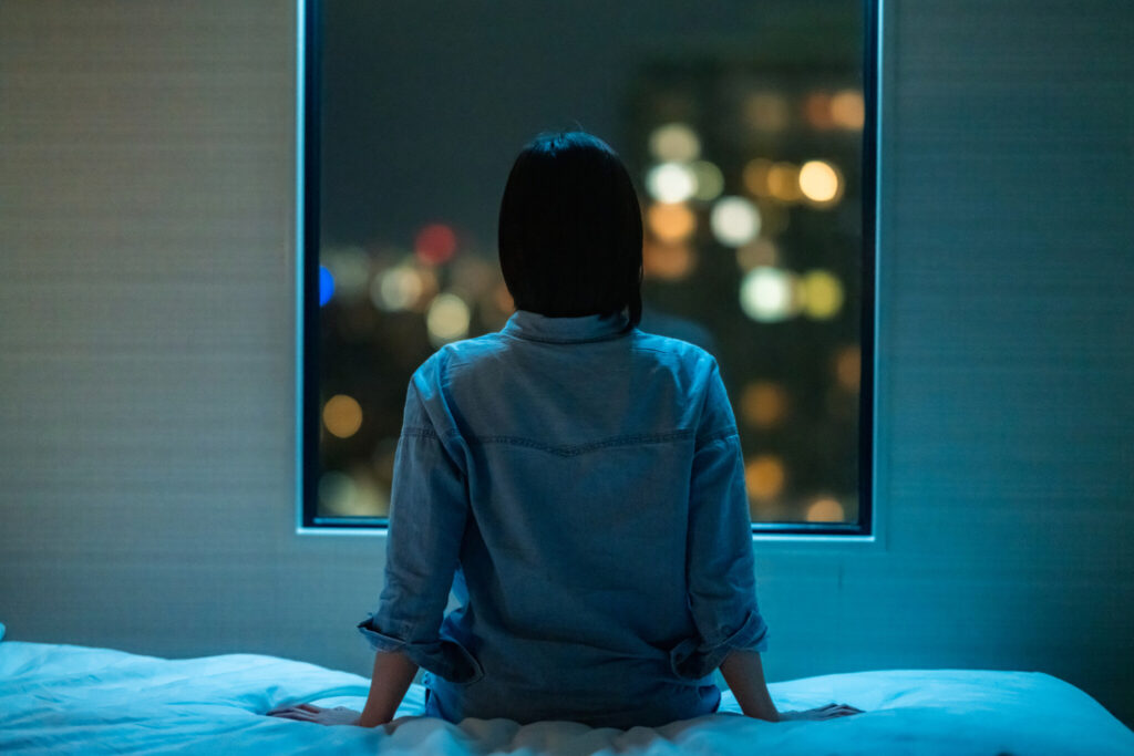 Rear view of woman sitting alone on bed in room and looking through window at night