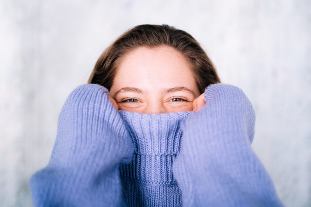 Beautiful young woman is covering face with violet sweater collar against gray wall background. She has short hair and gray eyes. Close-up portrait. Concept of natural beauty