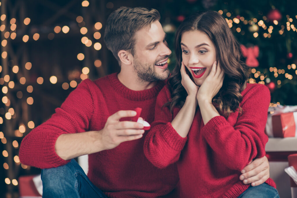 Photo of couple in decorated garland lights room guy giving lady unexpected engage ring box waiting answer sitting cozy near x-mas tree indoors wear red sweaters