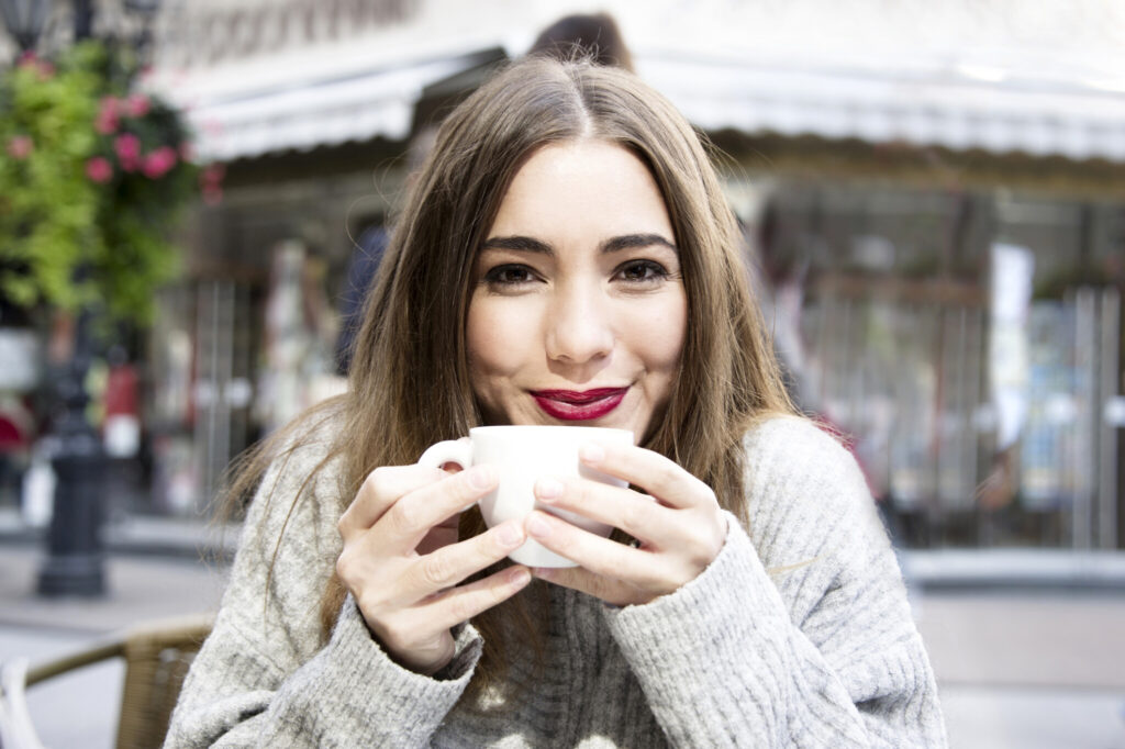 Attractive young woman is having a coffee on a street with street life background in autumn time.