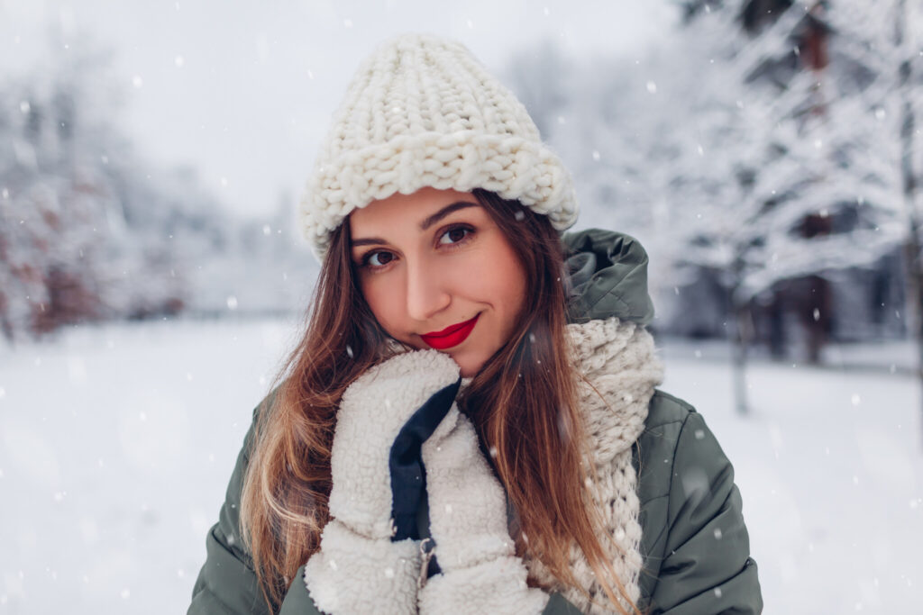 Close up portrait of happy young woman in snowy winter park wearing warm knitted clothes and red festive lipstick.