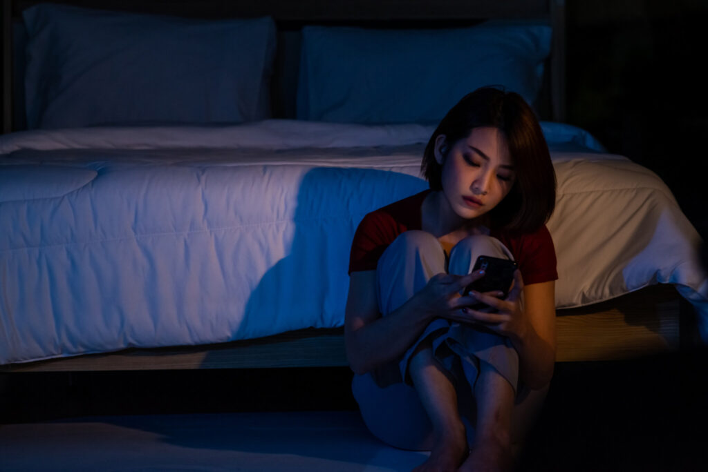 Depressed young beautiful Asian woman sitting alone on bedroom floor with looking at smartphone. Stressed insomnia sad woman having life problems. Mental health, healthcare and social issue concept.