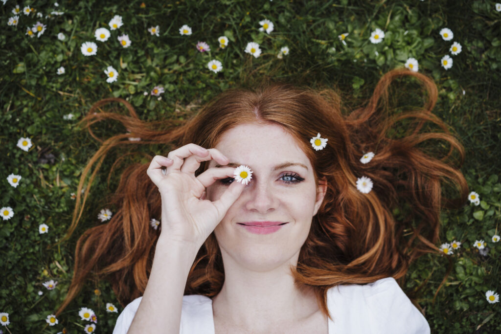 Smiling beautiful woman covering eye with daisy flower while lying on grass