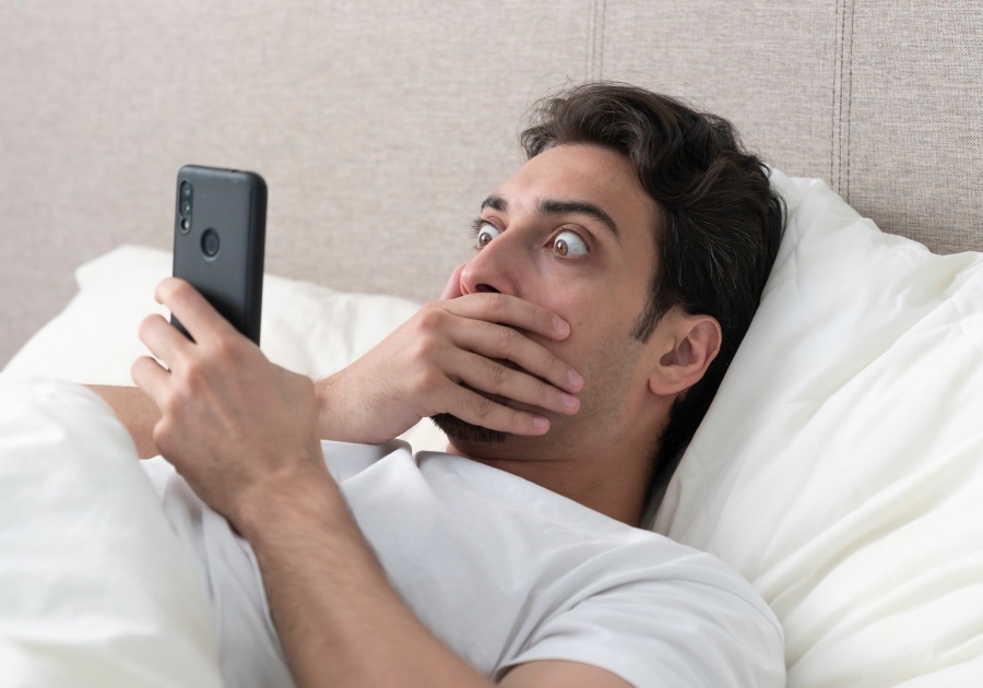 Surprised and shocked man lying on the bed and looking at smartphone at the morning time. In concept of bad news and checking up mobile after awakening